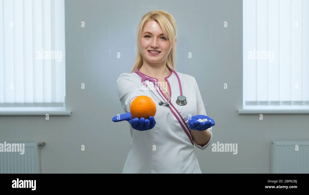 Caucasian young doctor in white medical coat, violet stethoscope holding a white pills with left hand and orange in rigth hand in blue gloves. Choice Stock Photo