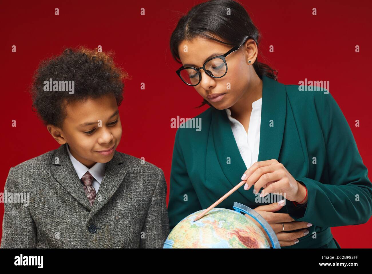 Close up portrait of young female teacher and cute African-American boy looking at geographical globe while standing against red background Stock Photo