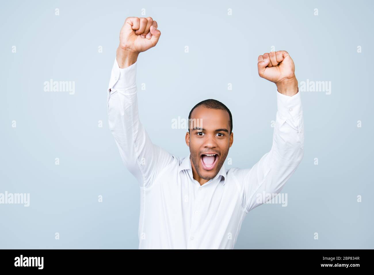 Attractive amazed african man, so shocked, extremely happy, with wide open mouth on pure grey background, in white shirt with raised hands Stock Photo