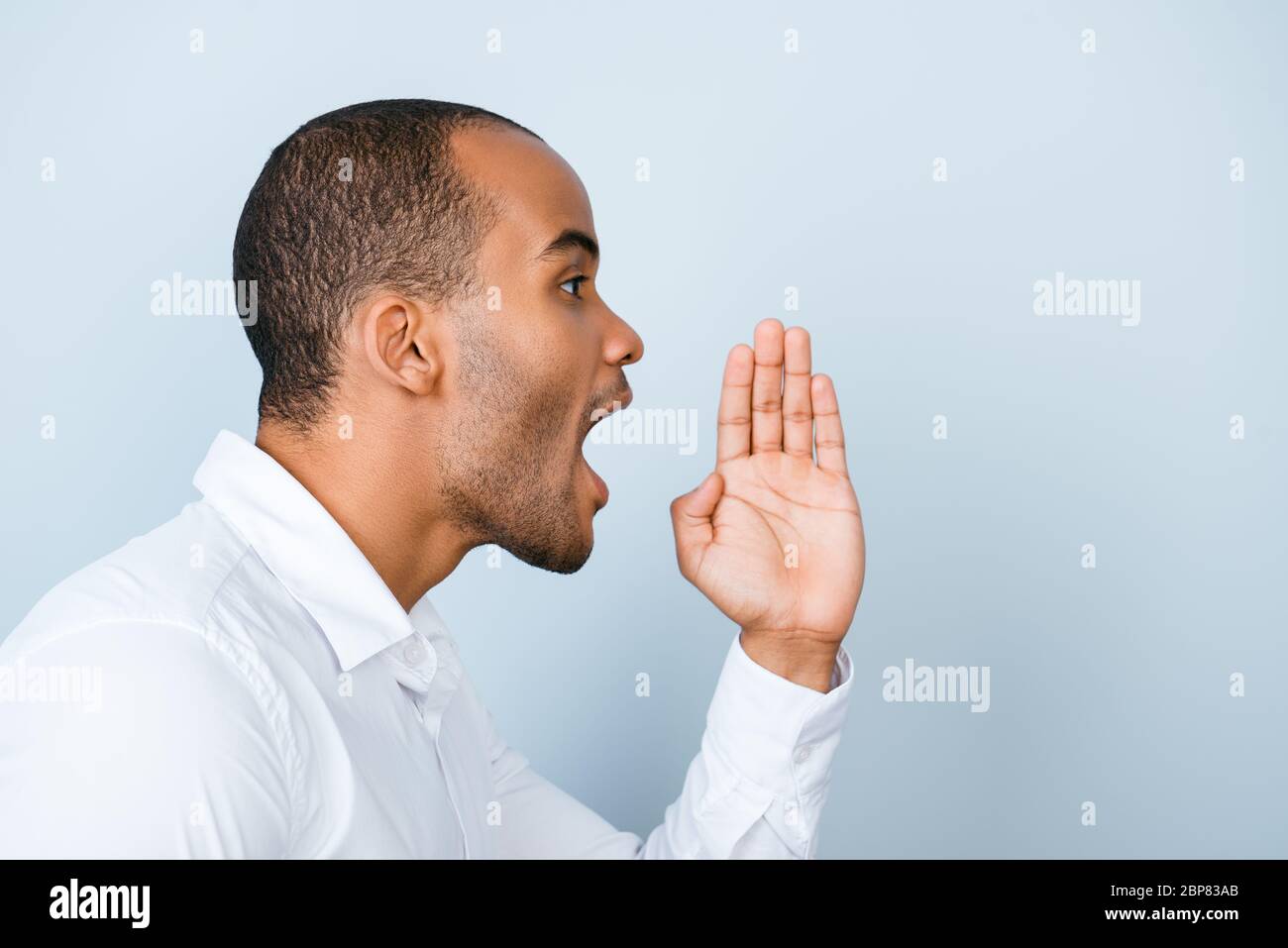 Attention! Cheerful young american mulatto man in white formal shirt is shouting and holding hand near her open mouth on light grey background Stock Photo