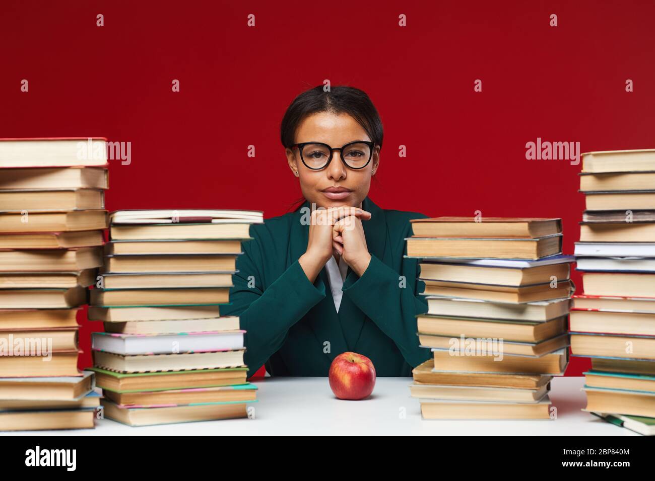 Front view portrait of young African-American woman sitting at desk between stacks of books and smiling at camera, copy space Stock Photo