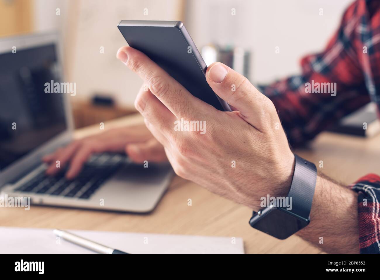 Using smart phone and laptop computer in home office, close up of male hands using modern electronics for telecommuting, selective focus Stock Photo