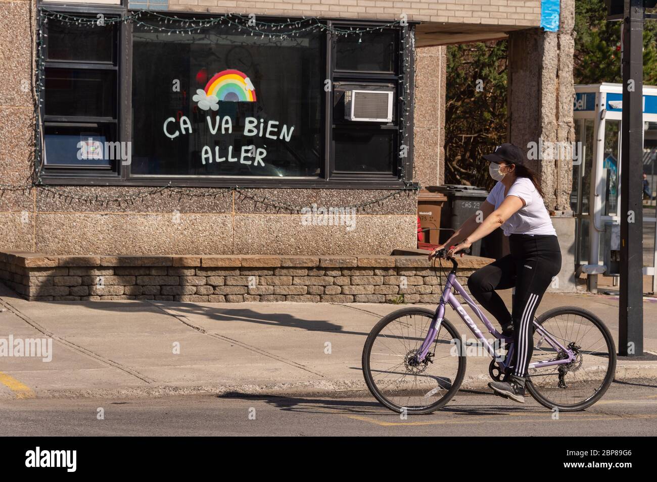 Montreal, CA - 17 May 2020: Young woman with face mask for protection from COVID-19 riding a bike in front of rainbow drawing on Masson street. Stock Photo