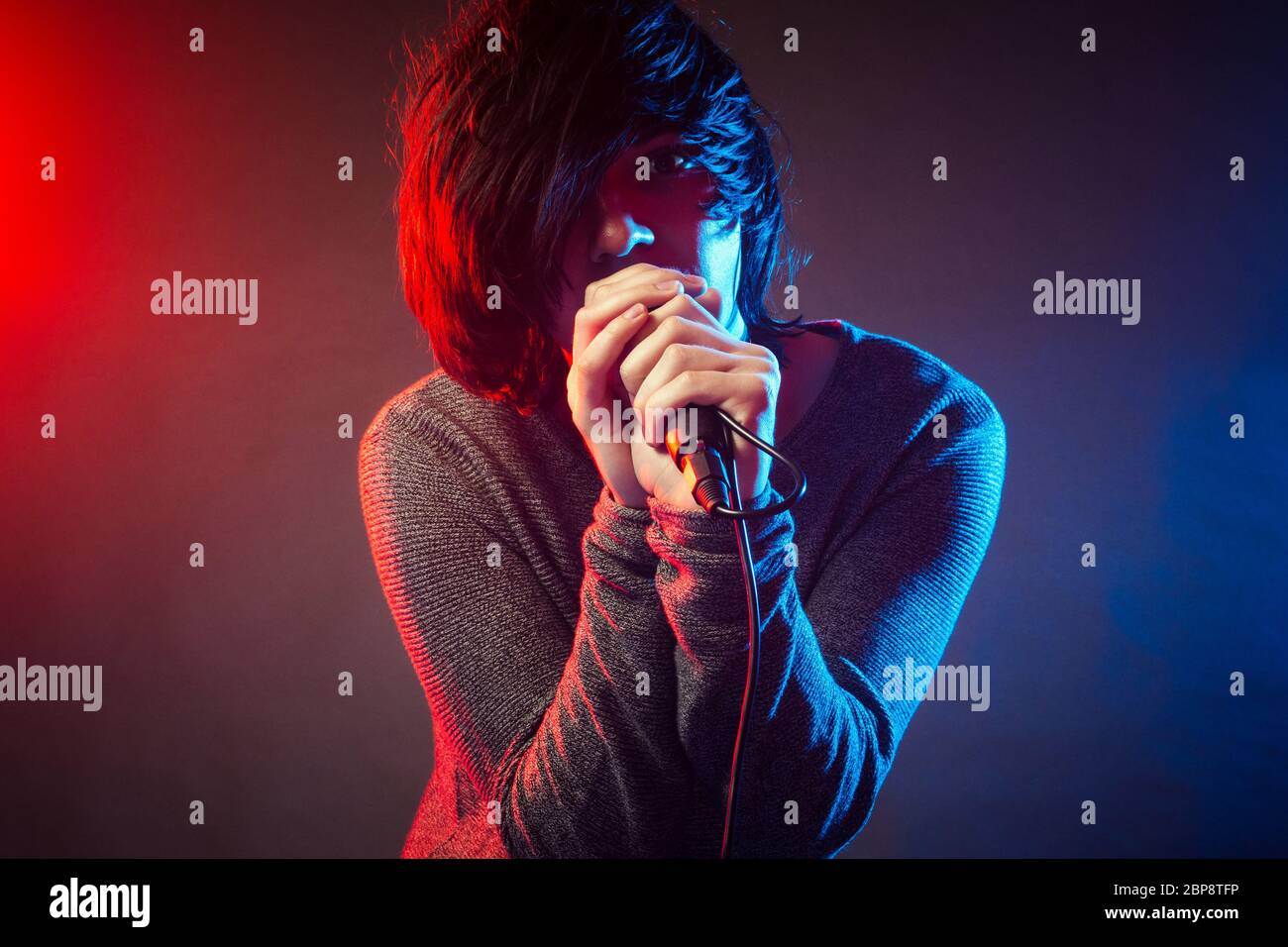 The young singer or vocalist in emo style is singing on concert on background of red and blue concert lights. Stock Photo