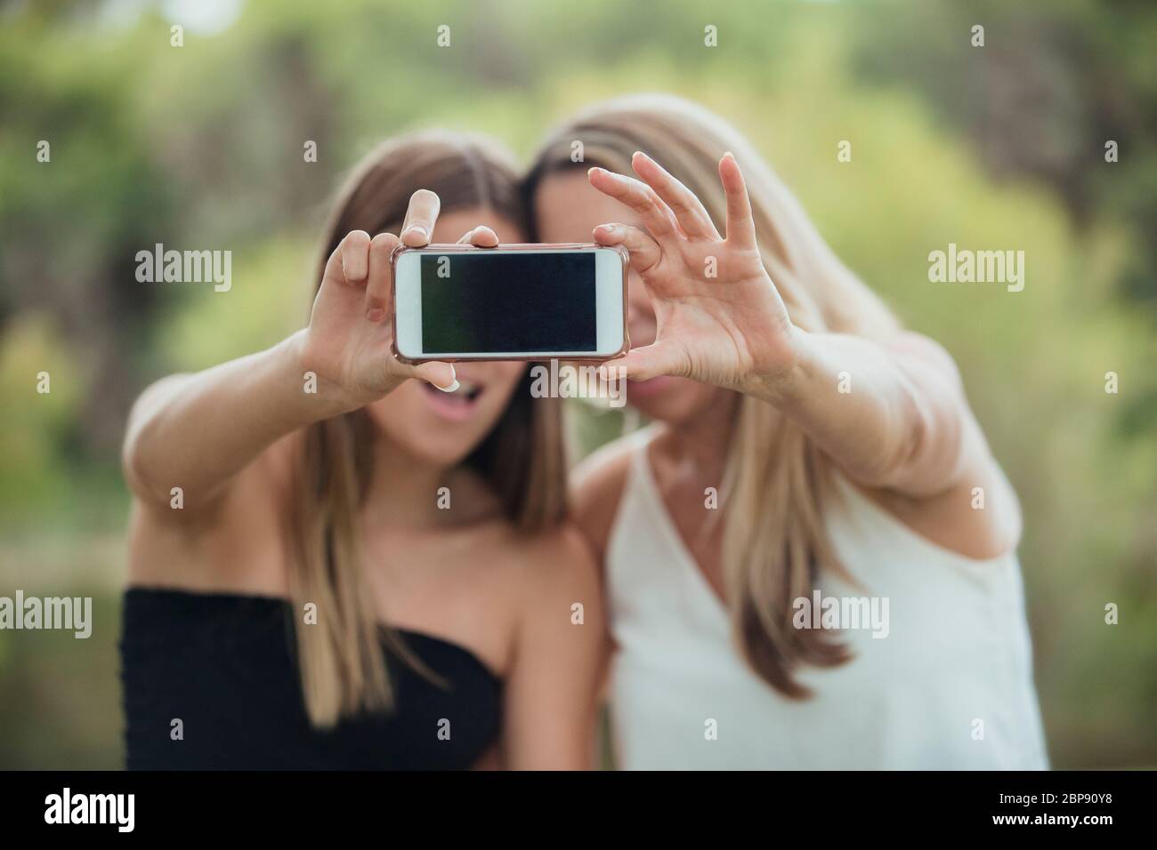 A mother and daughter are standing side by side outdoors capturing a selfie together. Stock Photo