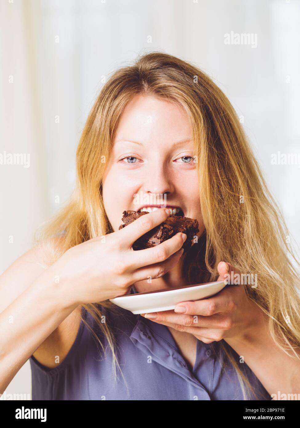 Photo of a beautiful blond woman in her early thirties with log blond hair eating a large piece of brownie or cake. Stock Photo