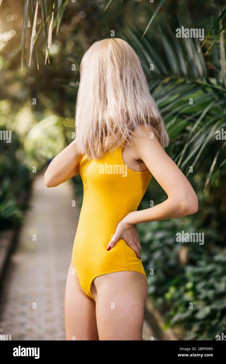 portrait of young beautiful blonde in yellow swimsuit in garden with tropical plants, Profile, face hidden. Slender caucasian woman touching the branc Stock Photo