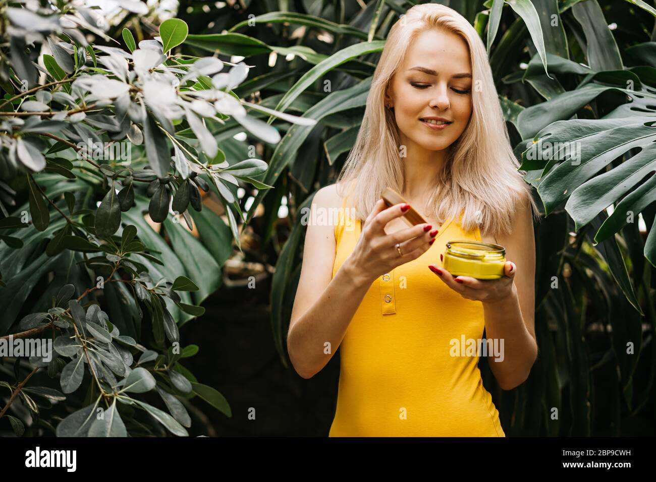 Beautiful caucasian woman with healthy smooth clean skin holding jar of body cream, sunblock, smelling its scent with closed eyes, posing in rainfores Stock Photo
