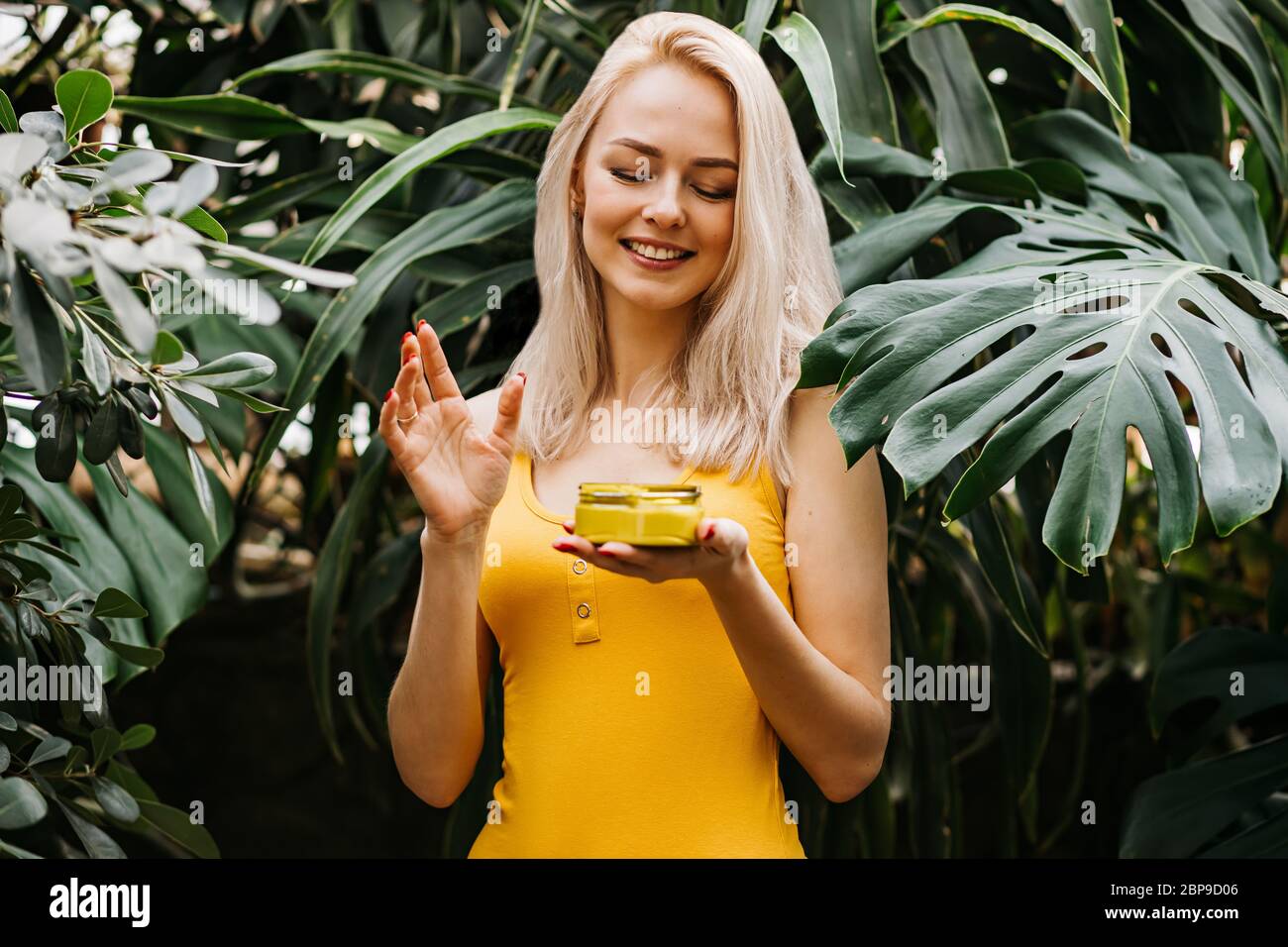 Beautiful caucasian blonde woman with healthy smooth clean skin holding jar of body cream, applying sunscreen, standing on tropical green leaves backg Stock Photo