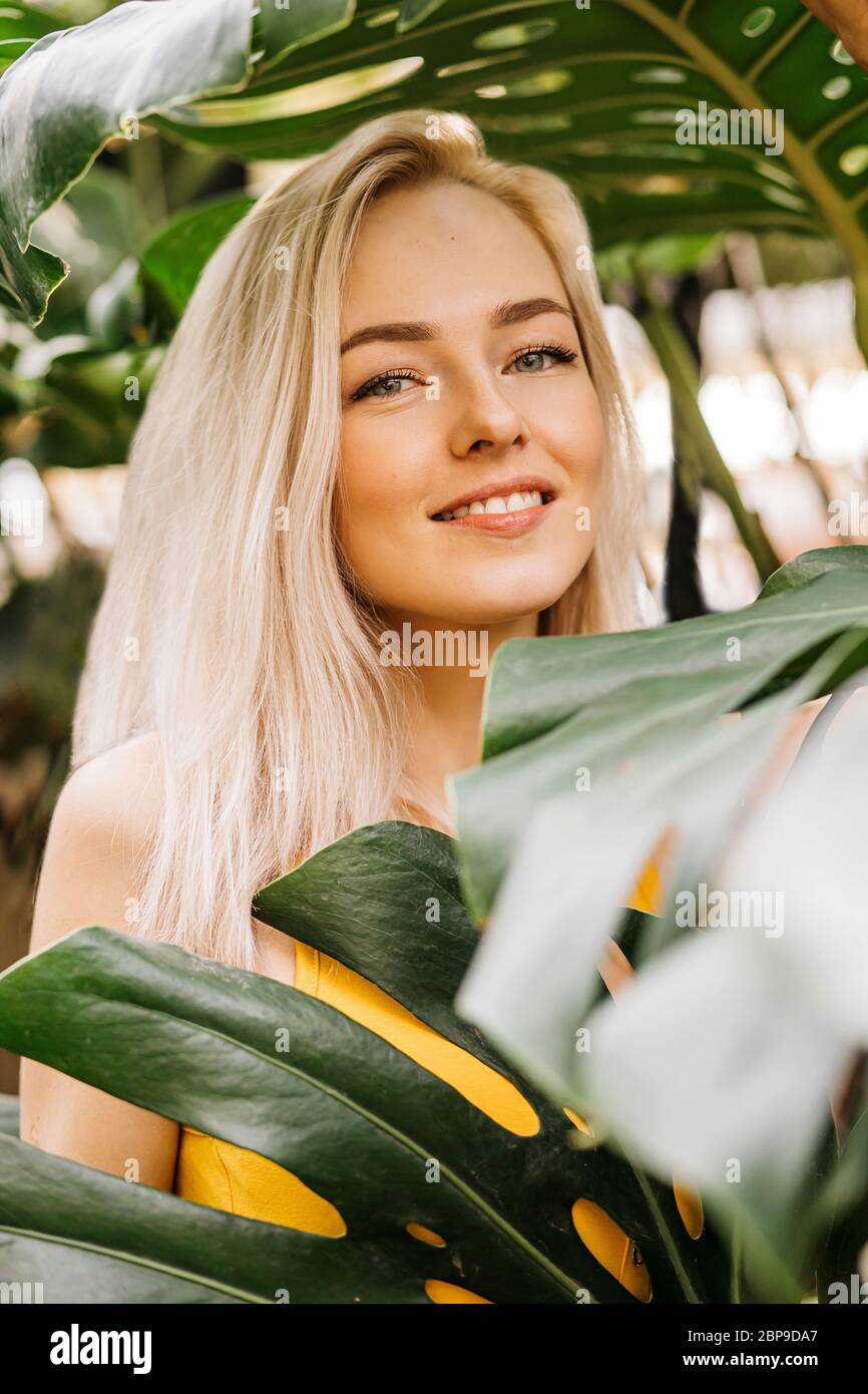 Closeup vertical shot of good-looking charming young blonde woman standing among green tropical plants. Attractive lady with natural beauty posing in Stock Photo