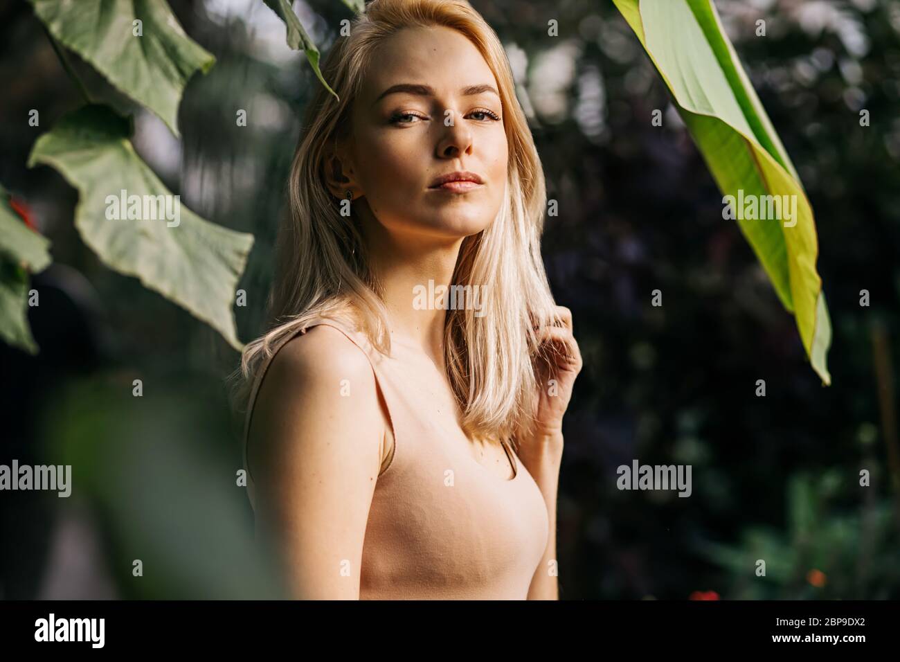 Portrait of beautiful young woman in forest . Lovely caucasian girl with natural makeup and blonde hair standing among exotic plants, looking into dis Stock Photo