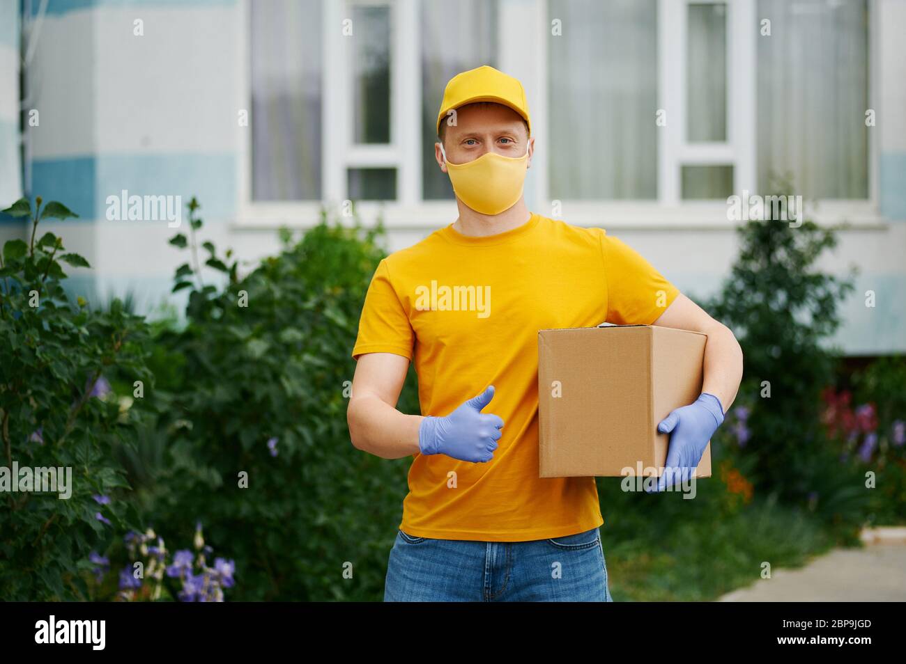Delivery Man employee in yellow uniform cap, t-shirt, face mask and gloves holds a cardboard box package and shows thumbs up . Safety delivery quarant Stock Photo