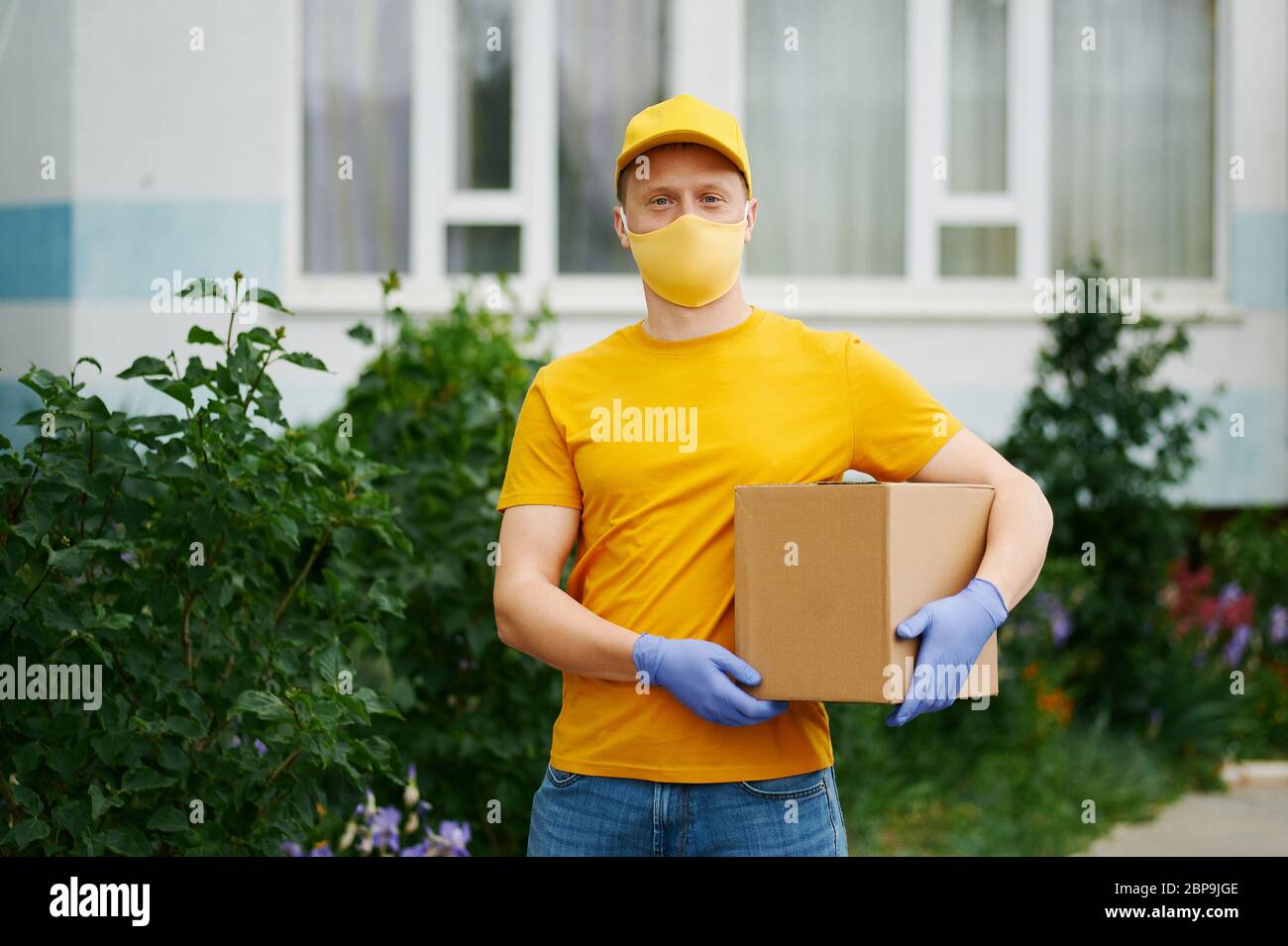 Delivery Man employee in yellow uniform cap, t-shirt, face mask and gloves holds a cardboard box package. Safety delivery quarantine service in covid- Stock Photo