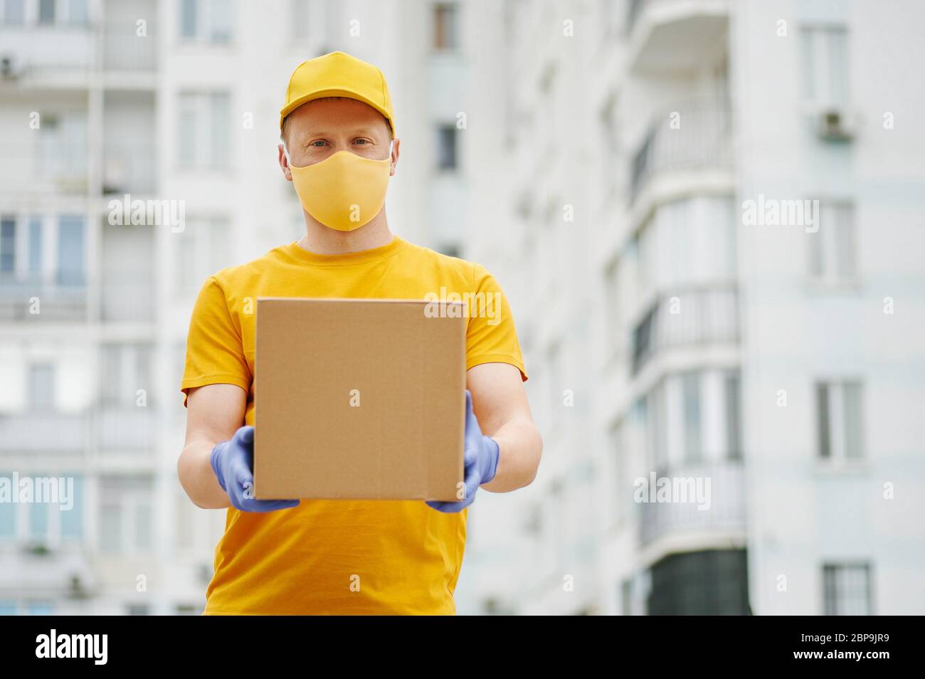Delivery Man employee in yellow uniform cap, t-shirt, face mask and gloves holds a cardboard box package on building backdrop. Safety delivery quarant Stock Photo