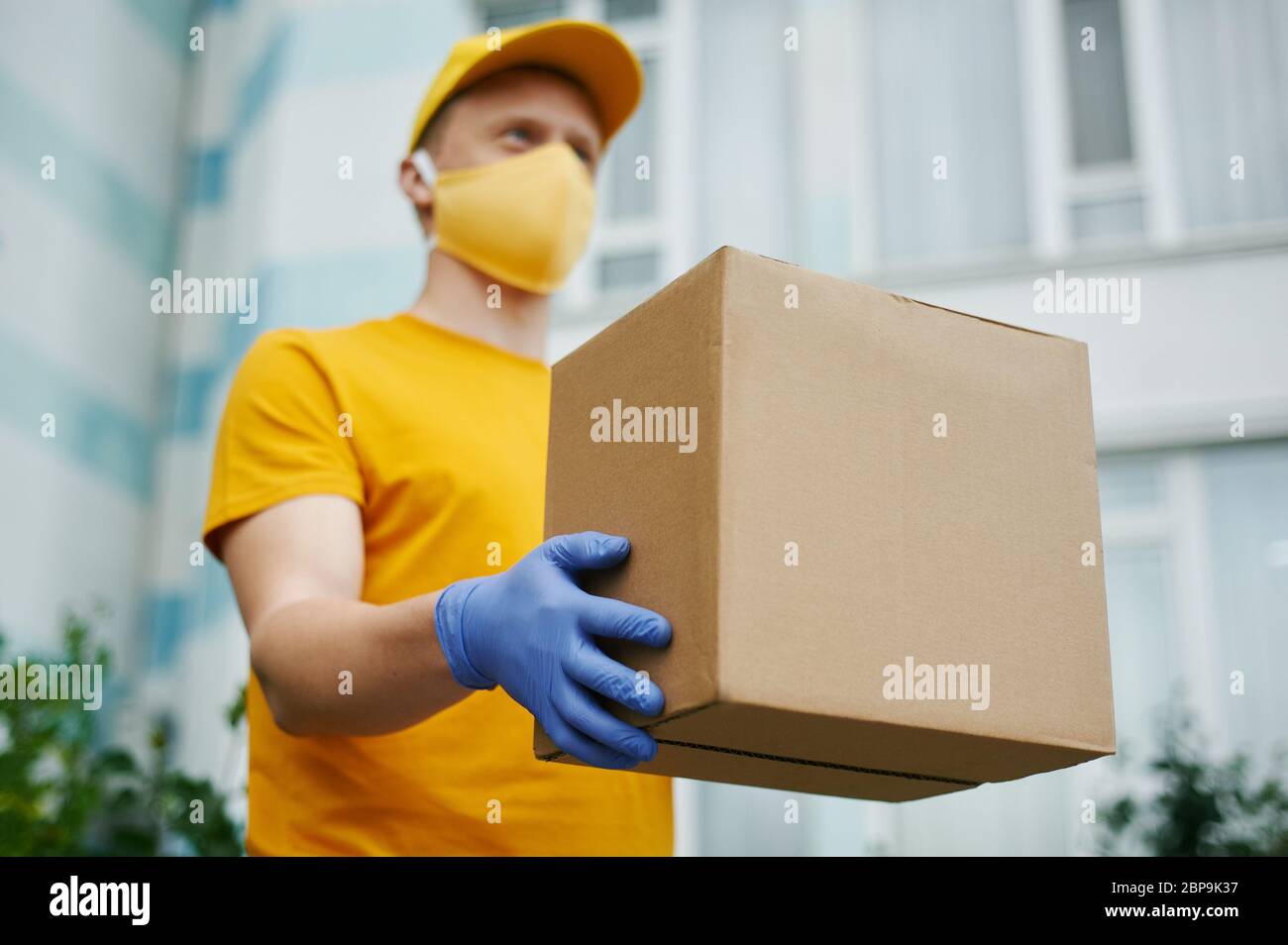Delivery Man worker in yellow uniform cap, t-shirt, face mask and gloves holds a cardboard box package on building backdrop. Safety delivery quarantin Stock Photo