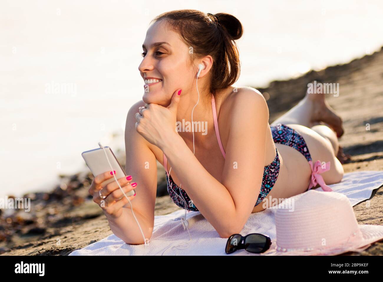 Attractive smiling woman with earphones in bikini is lying down, sunbathing and listening music from mobile phone at summer. Stock Photo