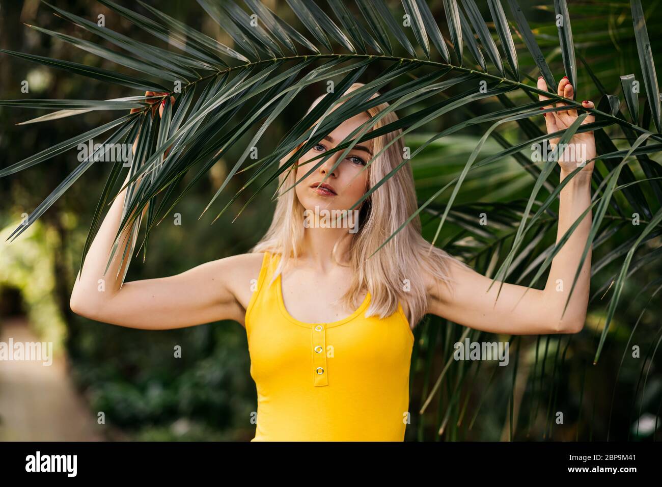 Closeup horizontal portrait of a beautiful young blonde woman in an exotic forest with green long leaves of a palm tree. Summer, vacation, skin care c Stock Photo