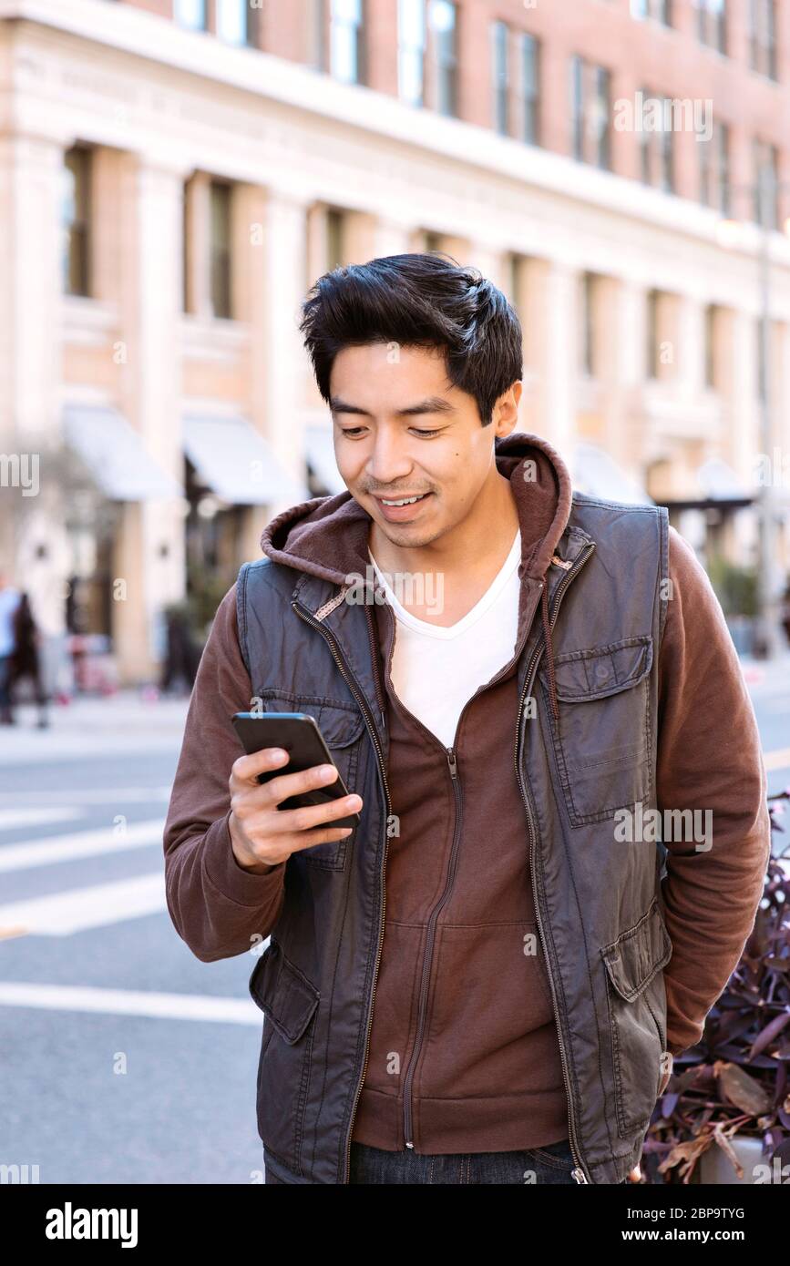 Millennial man looks at smart phone in the city - Using apps - Casual - Outdoors - Sunny Day Stock Photo