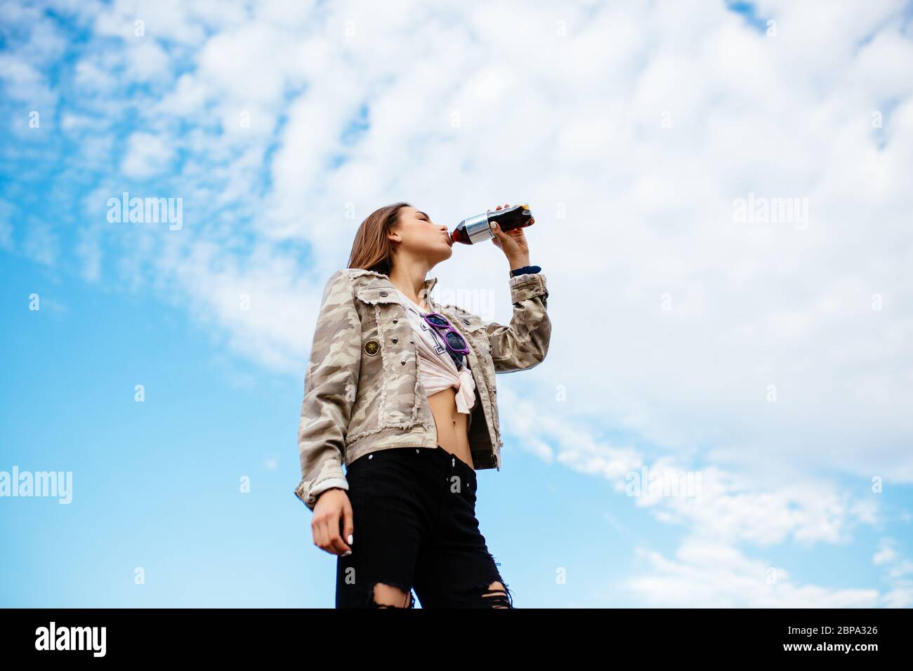 Trendy charming girl in fashionable clothes drinking a beverage from bottle, standing on the background of sky. Stock Photo