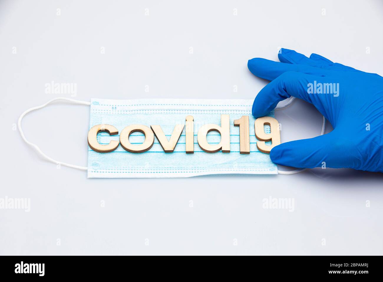 hand with a blue surgical glove holding the last part of the word covid19 formed with letters placed on a surgical mask isolated on a white background Stock Photo