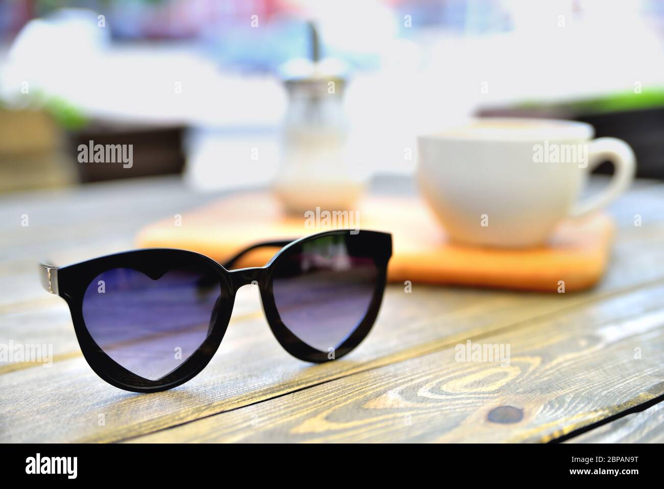 cup of coffee on a wooden stand is on the table in a cafe summer morning, sunglasses in the foreground, close-up Stock Photo