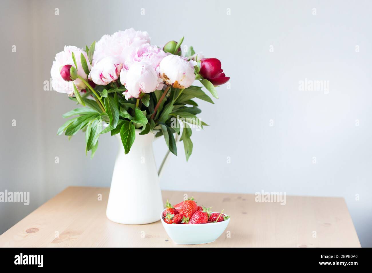 Bunch of Pink peonies in vase and strawberry on the wooden table . Flowers on a beige wooden table near the window. Home house interior. Beautiful Stock Photo