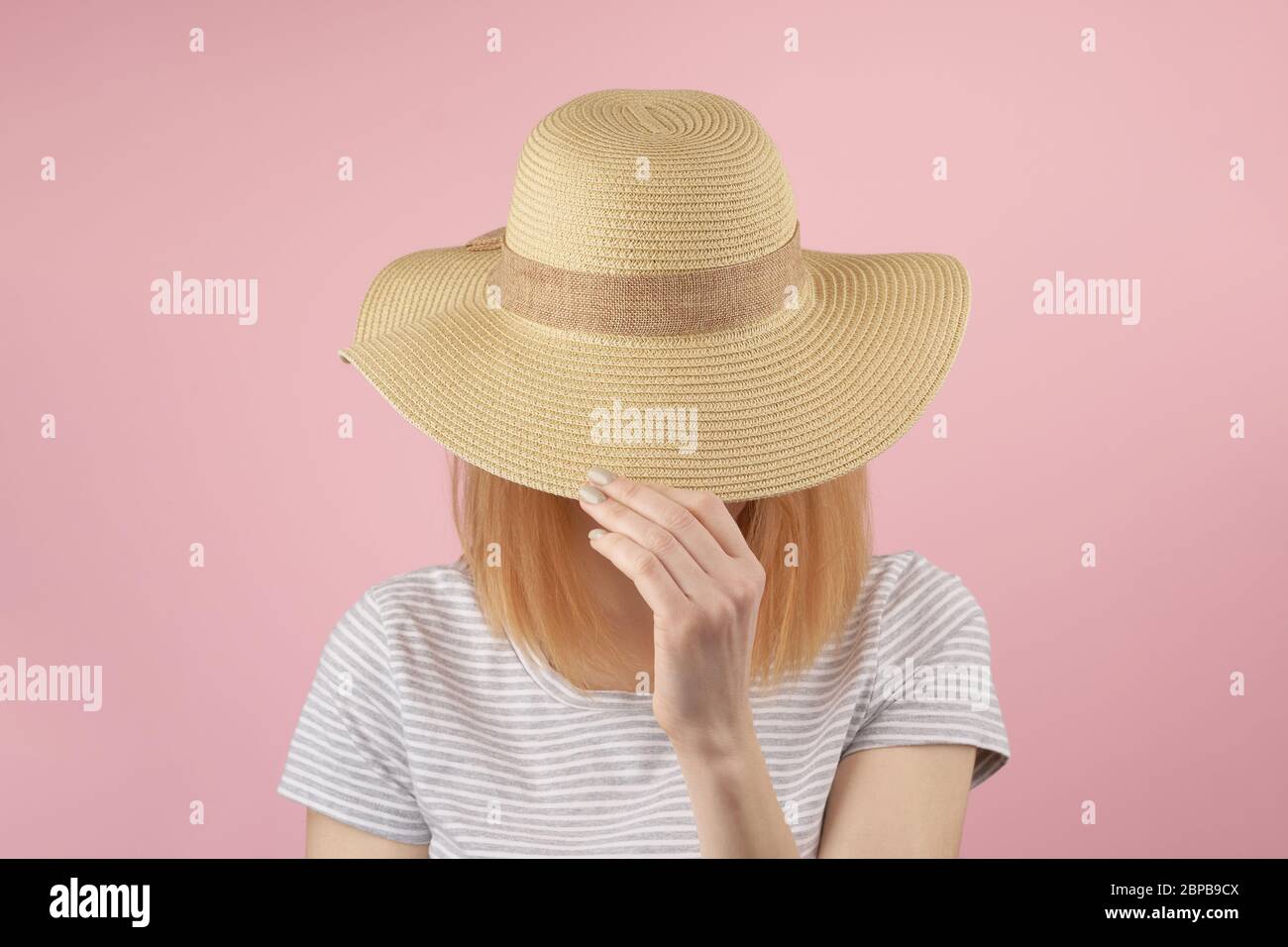 Redhair girl in gray tshirt hiding behind a straw hat as she pulls it down over her face with her hands. Frontal view upper body pink Stock Photo
