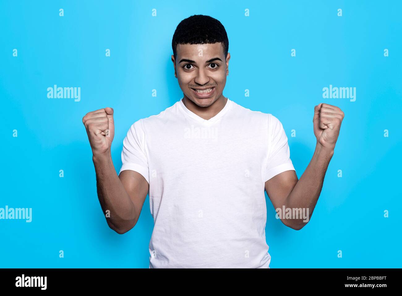 Overjoyed black male raises clenched fists, feels energized while gesturing happily against blue wall Stock Photo