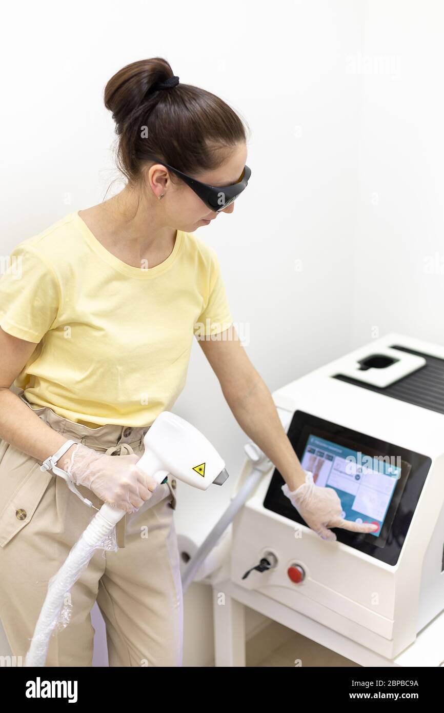 A young brunette girl cosmetologist selects the settings for laser hair removal and depilation. Vertical studio shot of the device. On hands are prote Stock Photo
