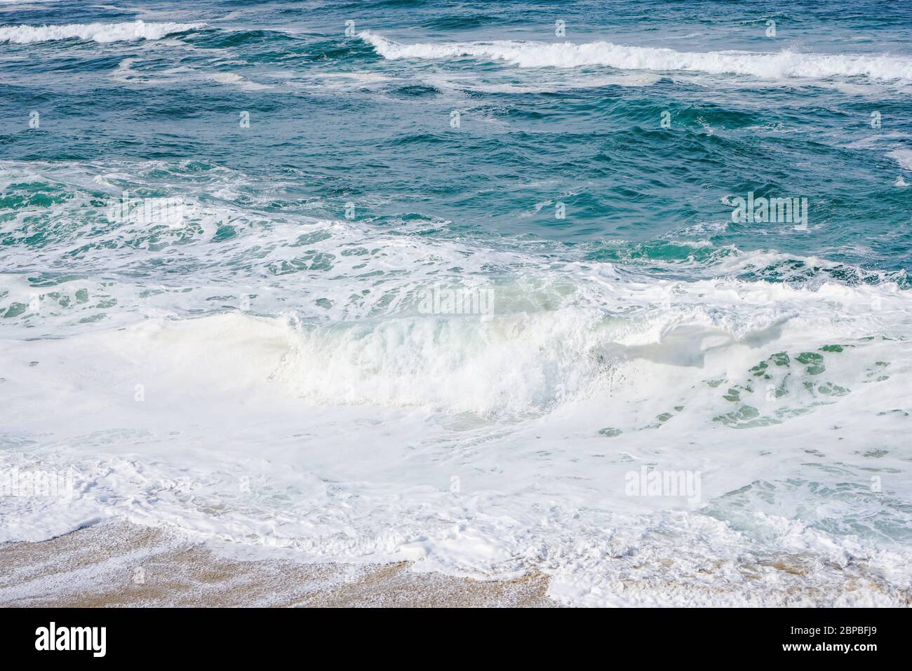 Turquoise ocean water with small white foam waves gently crashing into golden sand beach Stock Photo