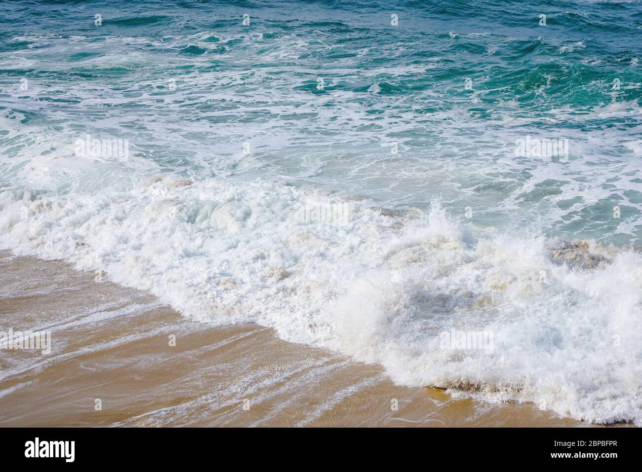 Turquoise ocean water with small white foam waves gently crashing into golden sand beach Stock Photo