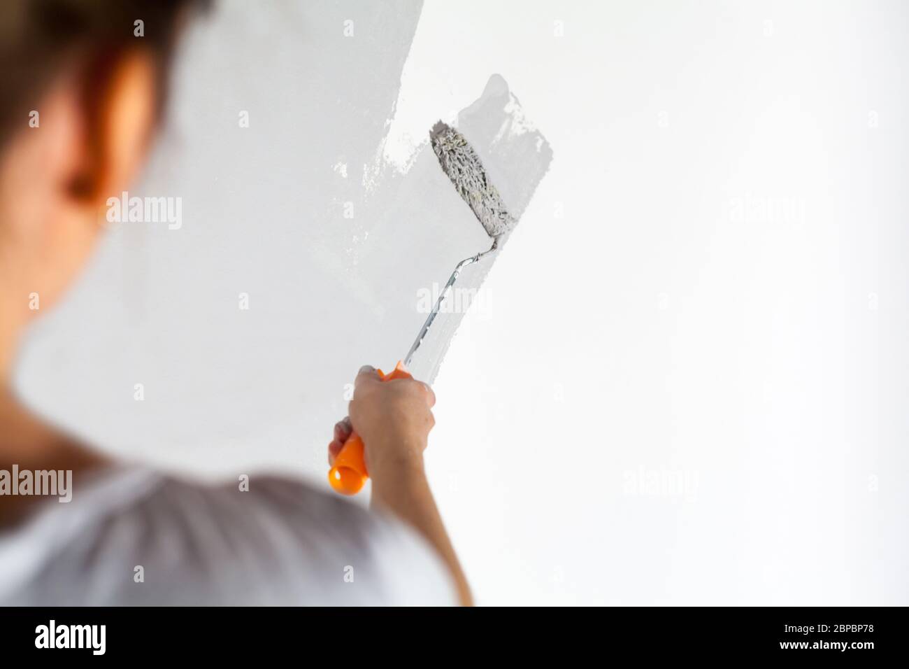 Craftswoman with small paint roller paints white wall with silver-gray paint painting company training teaching profession do it yourself tools Stock Photo