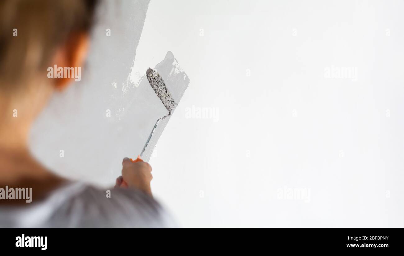 Craftswoman with small paint roller paints white wall with silver-gray paint painting company training teaching profession do it yourself tools Stock Photo