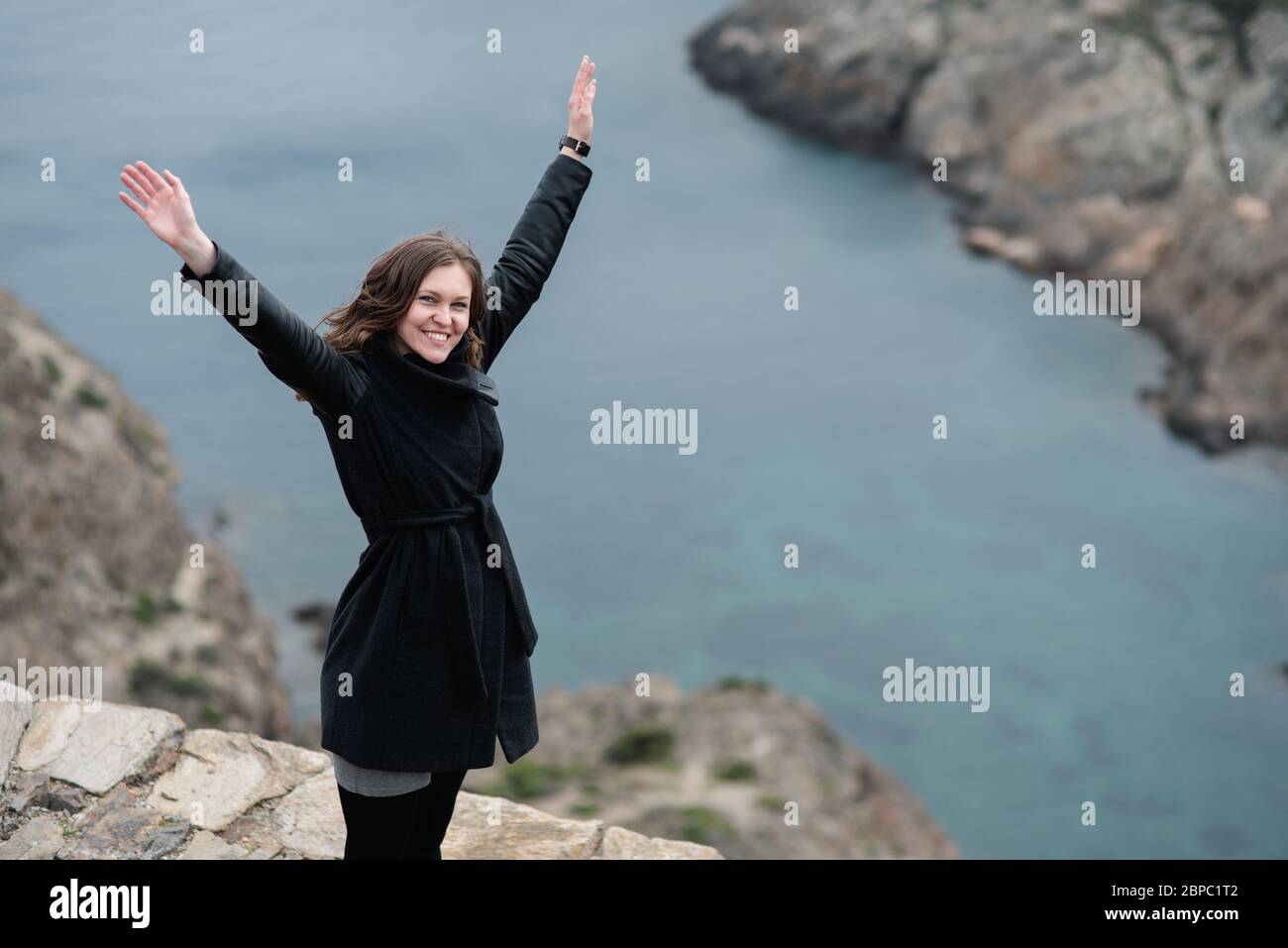 woman raises her arms to the sky as a sign of freedom on top of a cliff in front of the sea Stock Photo