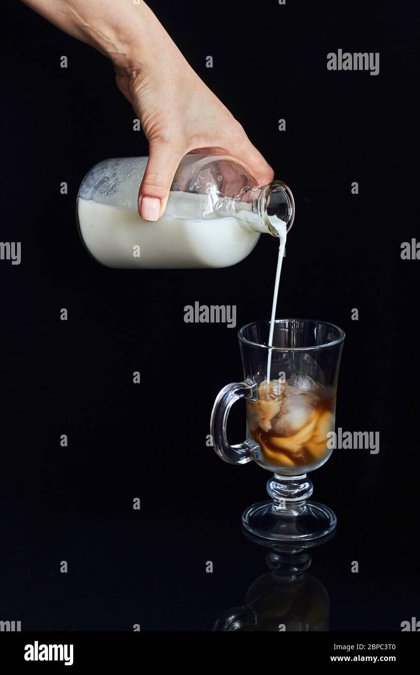 Female hand pours milk or cream from bottle into glass with ice coffee, cold coffee concept on black background Stock Photo