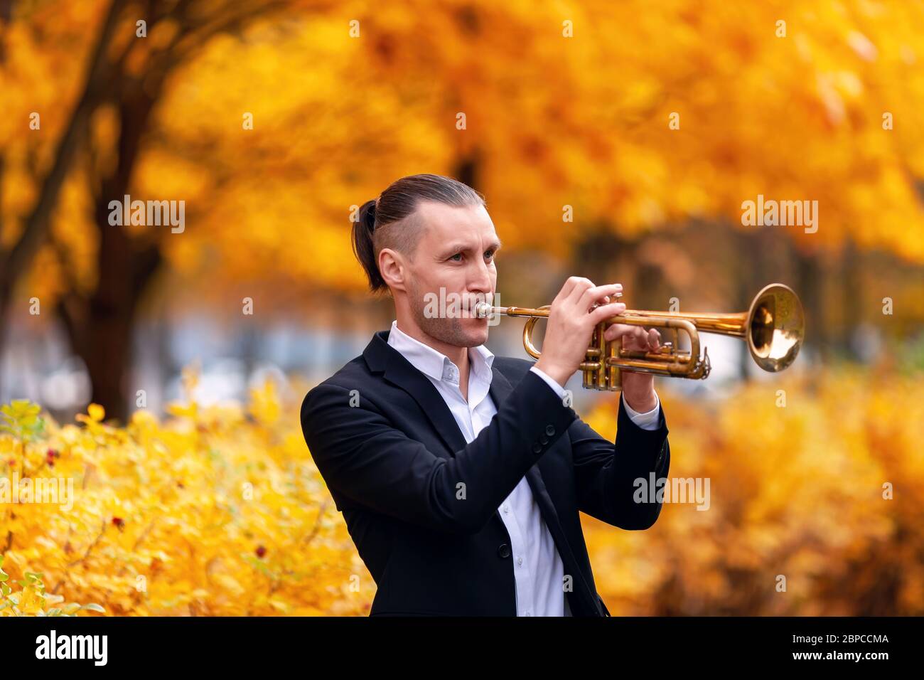 young European handsome trumpeter in formal clothes playing his musical instrument golden trumpet among trees with yellow leaves in autumn park Stock Photo