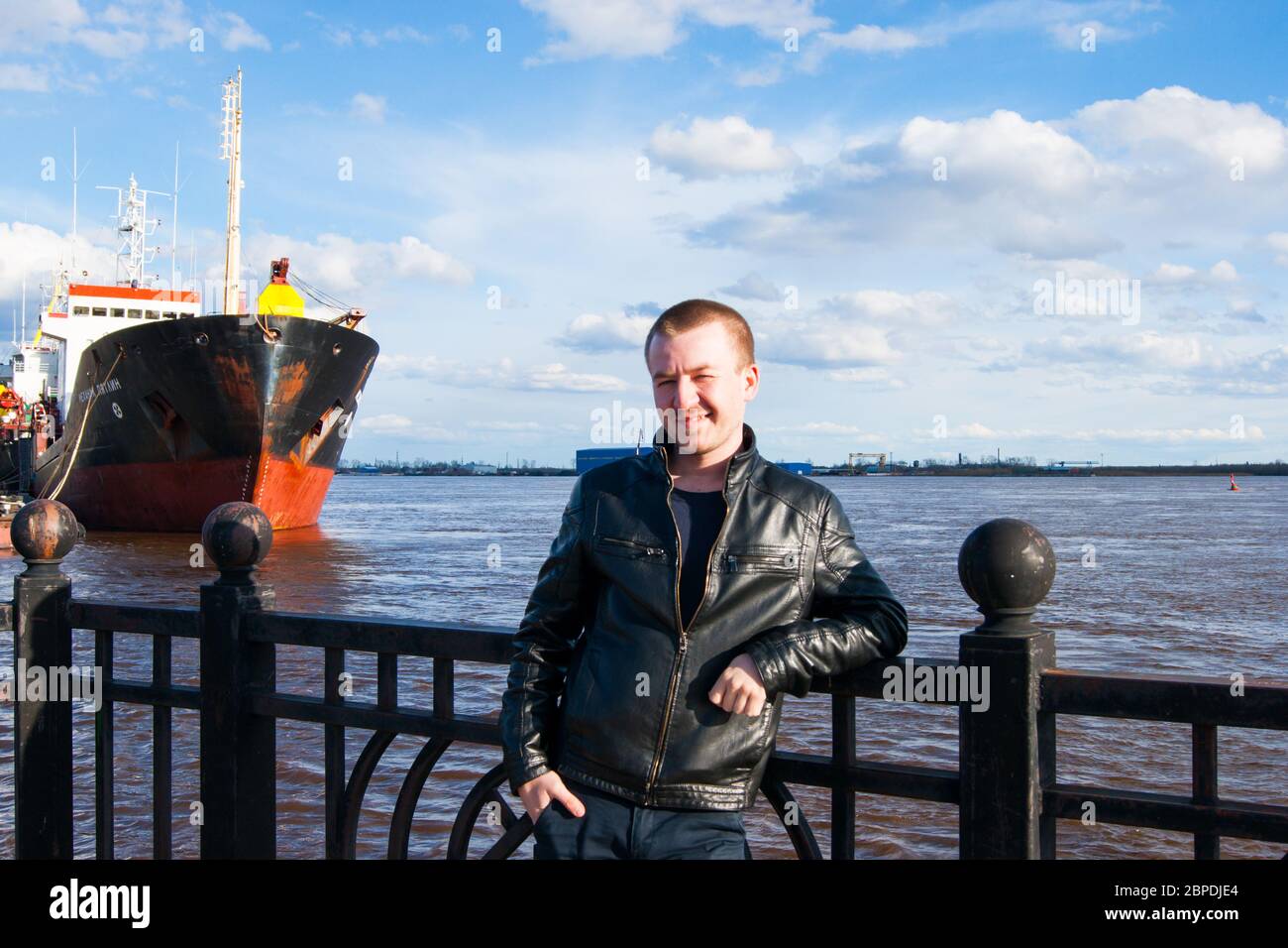 A young man - a blond Caucasian type stands on the embankment against the background of a river and ships, smiling. Spring, river, pier, river transport, spring, sky with clouds Stock Photo