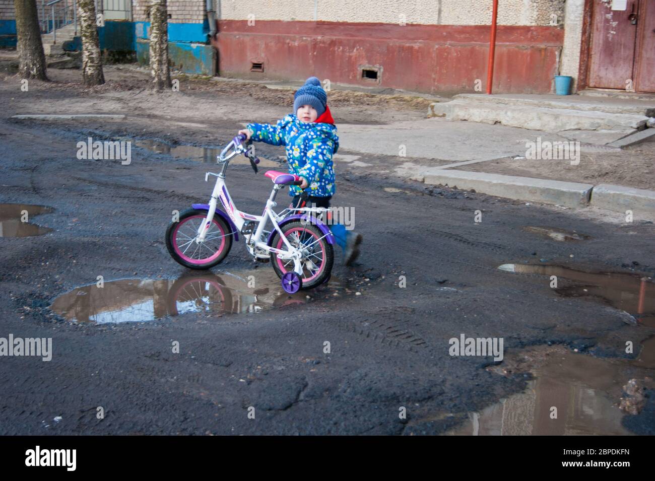 A child of three or four years walks with a two-wheeled bicycle on broken spring roads and puddles. Bad roads, asphalt, off-road, spring, walks Stock Photo