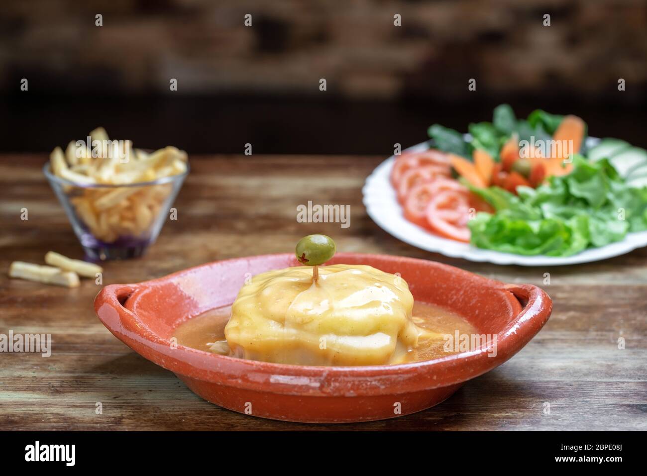 Portuguese francesine in a clay plate with salad and fried potatoes on a wooden background. Stock Photo