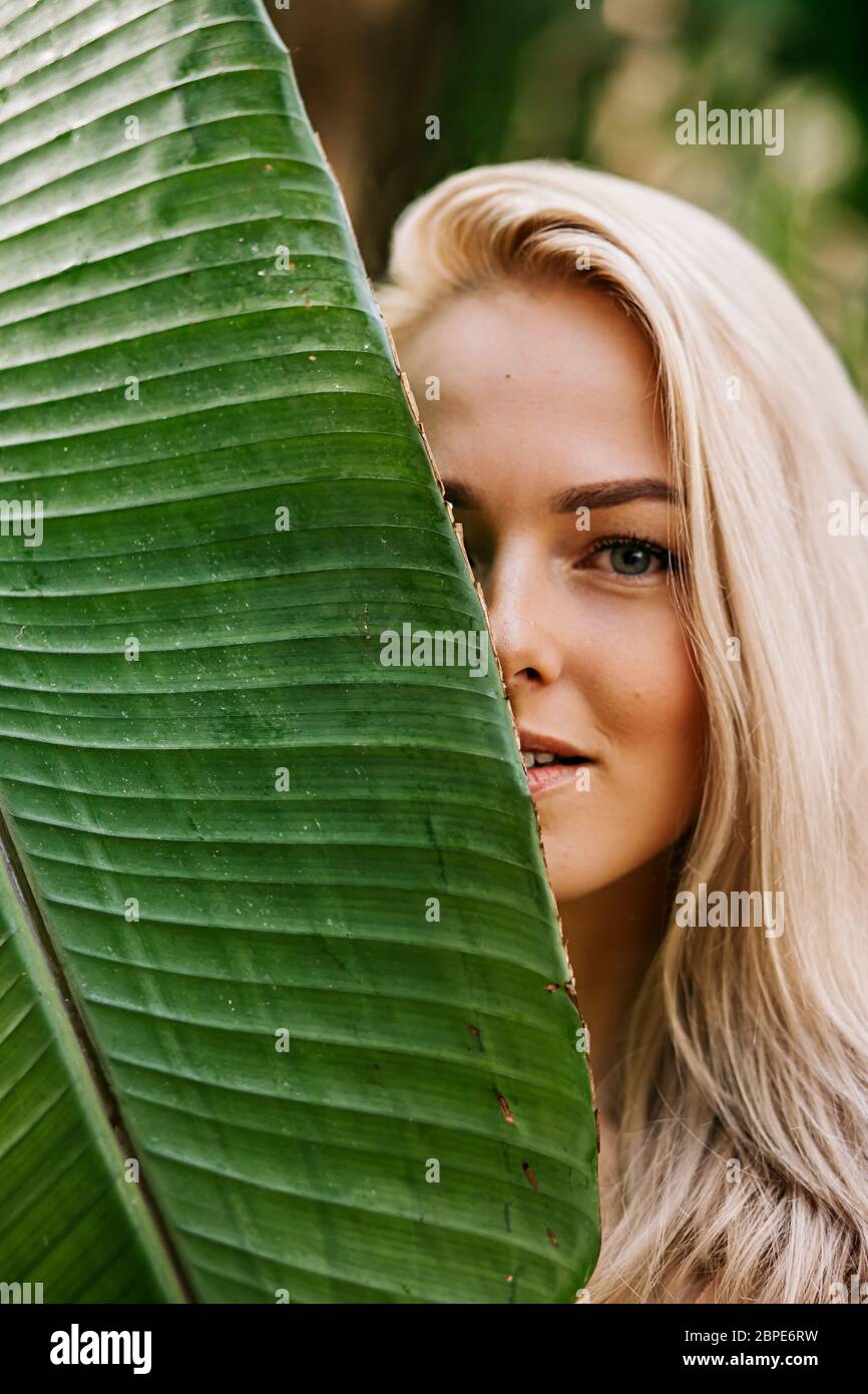 Closeup portrait of pretty caucasian lady with blonde hair, natural makeup, clean fresh skin, hiding half of face behind big tropical leaf. Skin care, Stock Photo