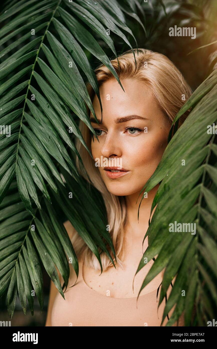 Portrait of happy lady with natural nude makeup, perfect skin, blonde hair. Beautiful woman hiding behind palm leaves and look down. Cosmetic, wellnes Stock Photo