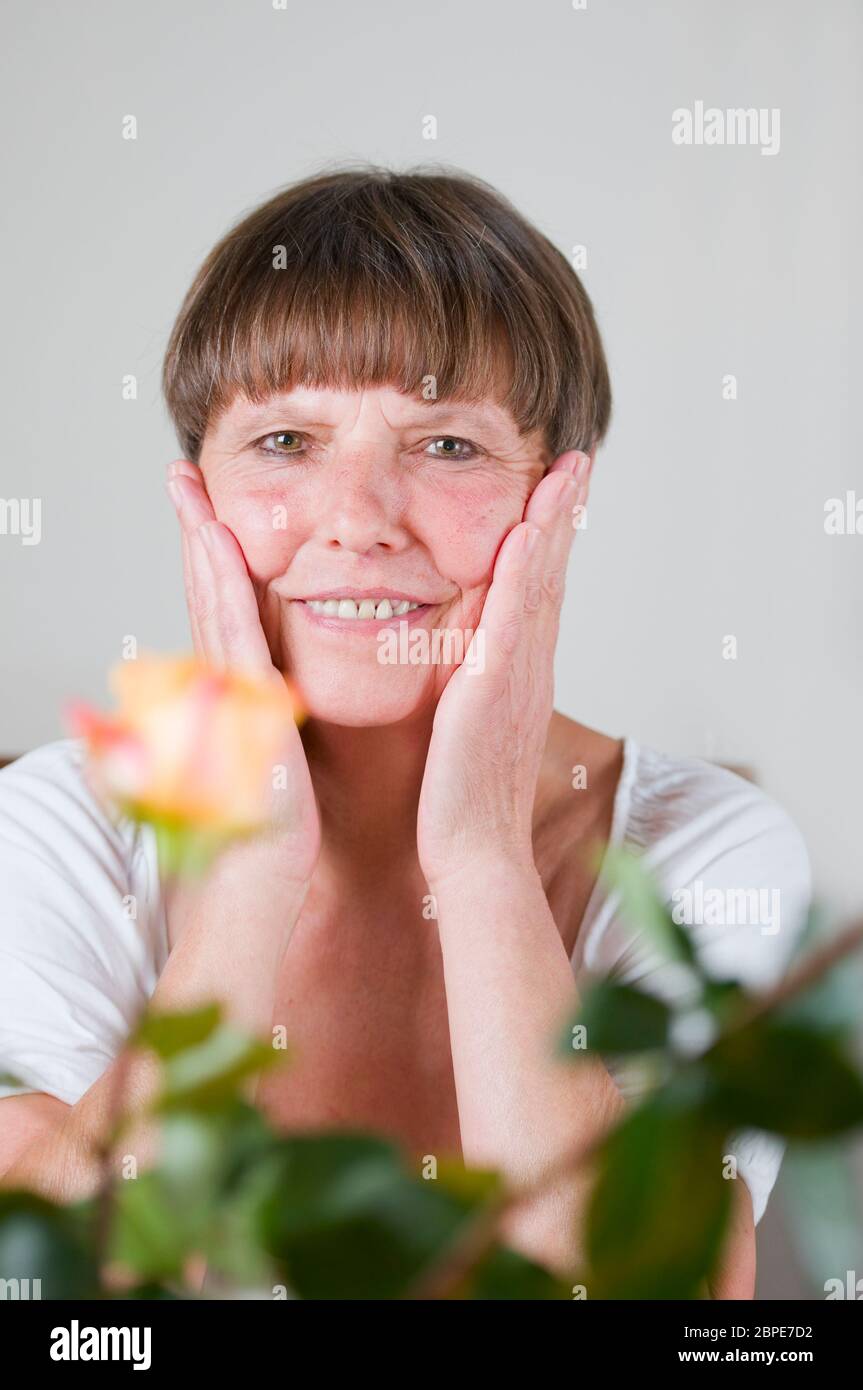 frontales Porträt einer älteren Frau hinter Blumen am Tisch sitzend sich die Hände an die Wangen haltend und in die Kamera lächelnd Stock Photo