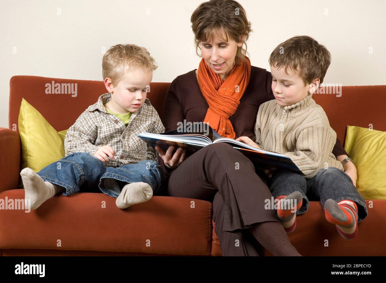 Mother and her two sons reading a book on a couch. Stock Photo