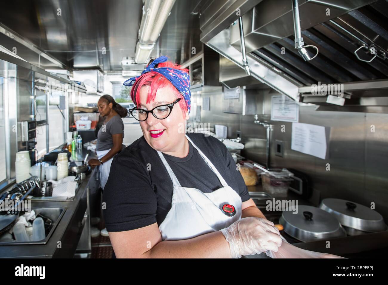 Pink haired chef puts on work gloves aboard busy food truck Stock Photo