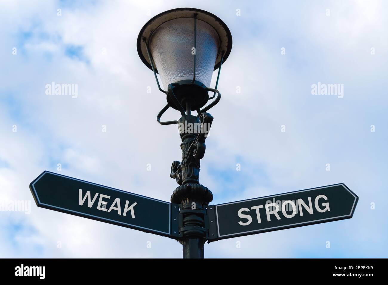 Street lighting pole with two opposite directional arrows over blue cloudy background. Weak versus Strong concept. Stock Photo