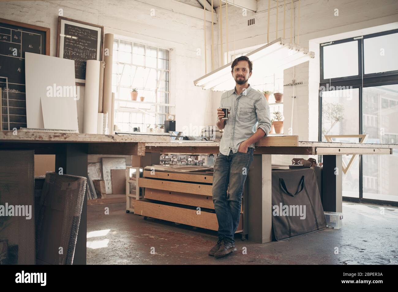 Full length portrait of a young male designer standing comfortably in his naturally lit workshop, holding his morning cup of coffee and smiling at the Stock Photo