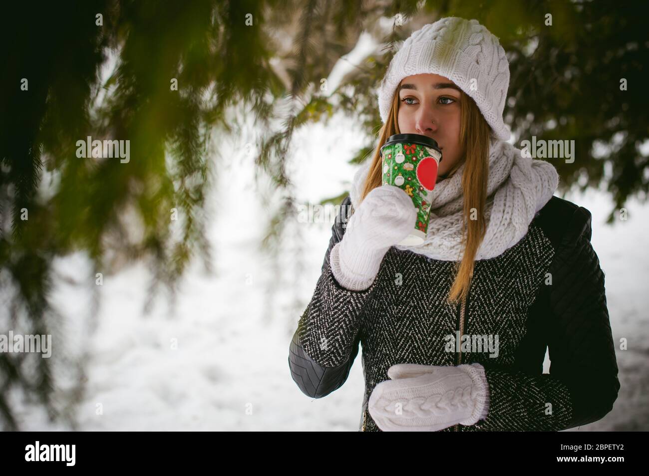 Portrait of cute woman in white scarf and hat knitted coat on outdoors background of snow and blurred fir branches in winter. girl holds a coffee cup Stock Photo