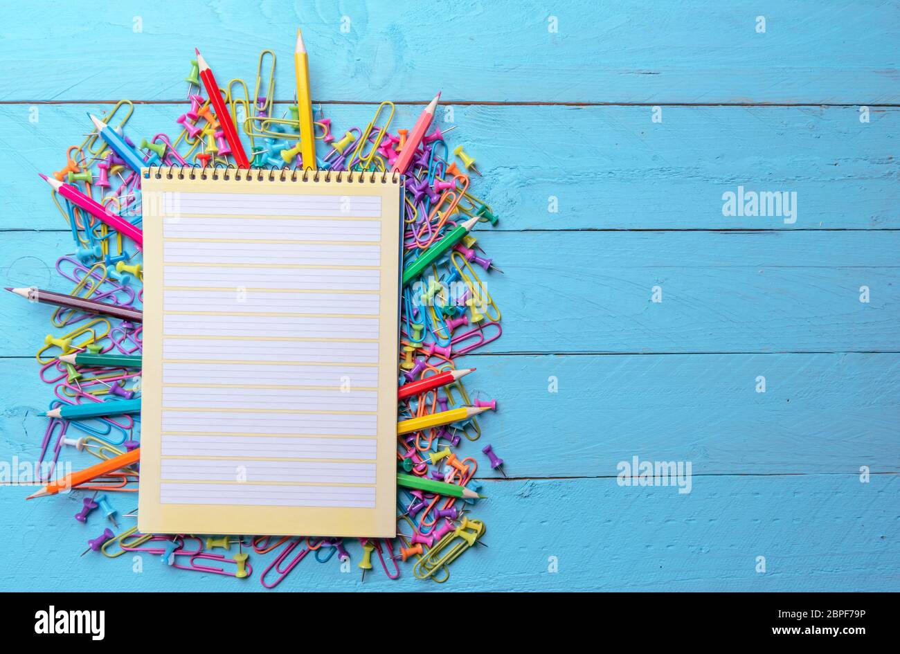 Blank page spiral notebook on a pile of office supplies, colored paper clamps, push pins and wooden crayons. Stock Photo