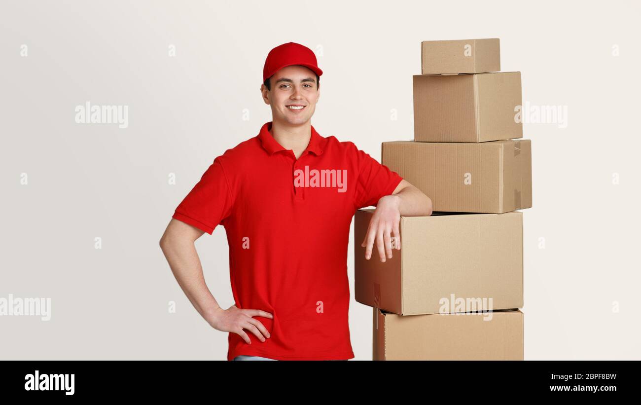 Smiling courier stands in storage warehouse and puts hand on boxes, studio shot Stock Photo