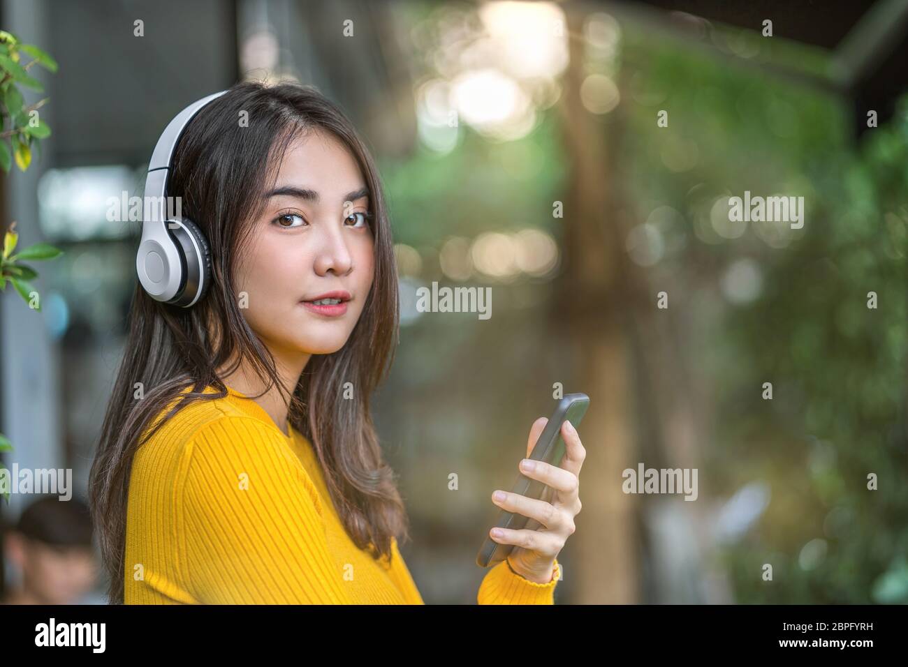 Happiness Asian smiling young woman wearing technology wireless headphones for listening the music via smart mobile phone infront of coffee shop or co Stock Photo